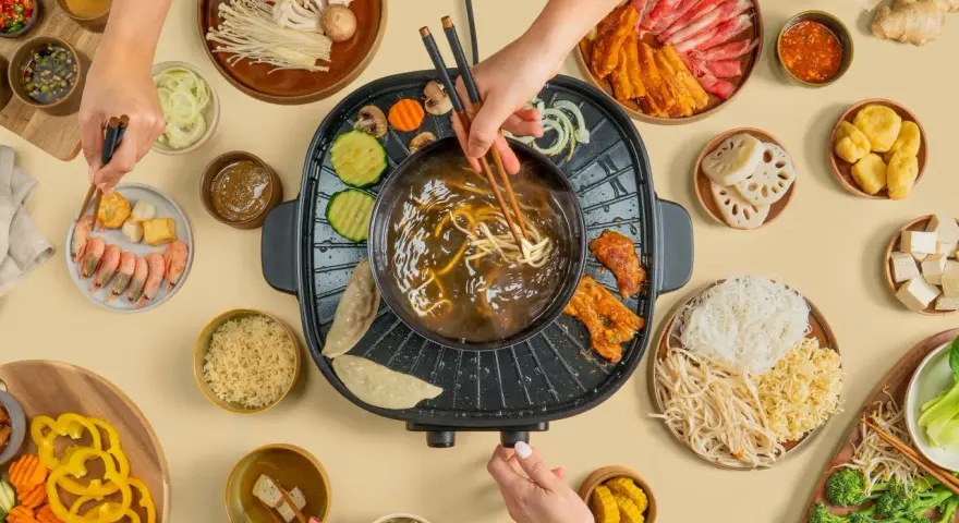 Hände greifen nach Zutaten für einen Hot Pot. Fleisch, Gemüse, Garnelen und Nudeln werden in die köchelnde Brühe getaucht.