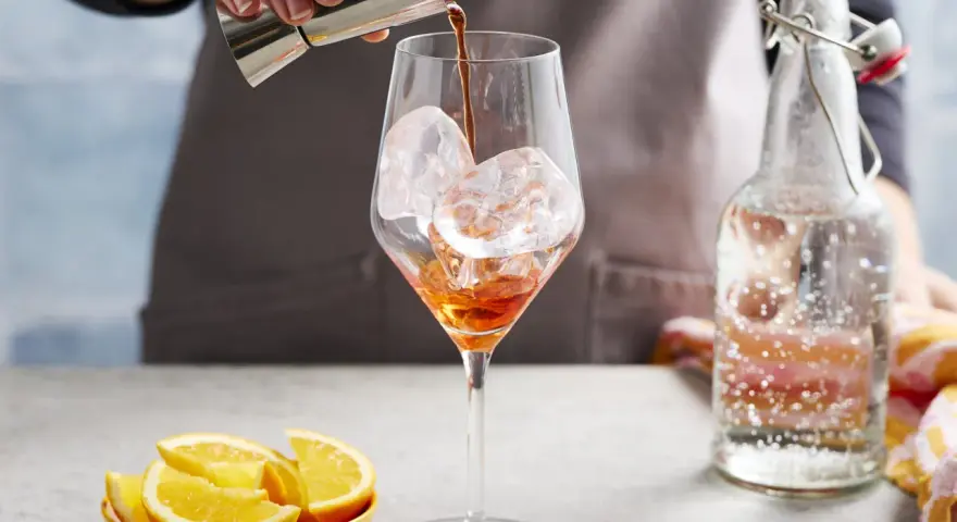 Zabawne przygotowanie Aperol Spritz Rezept Original. Likier wlewa się do kieliszka z lodem. Obok leżą pomarańcze i butelka prosecco.