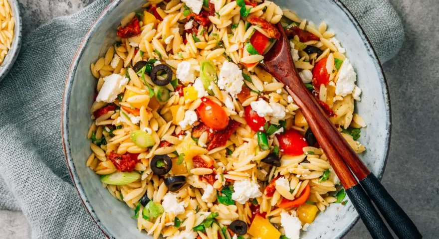 Bunter Kritharaki Salat mit Tomaten, Oliven, Feta und Kräutern, serviert in einer Schüssel mit Holzstäbchen.