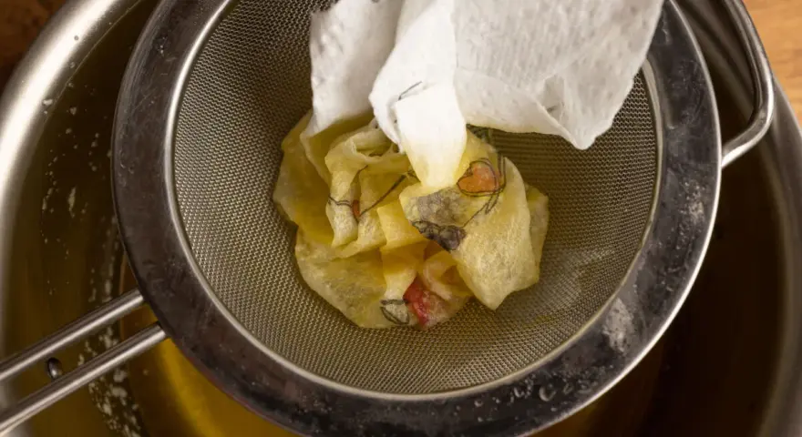 Ein Sieb hält ein Stück Papier mit Blumenmuster, das über einem Topf mit geschmolzener Butter liegt. Perfekt für raffinierte Butter Rezepte.