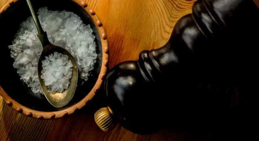 Fleur de sel, zarten Salzkristallen auf einem Löffel in einer Schale, neben einer dunklen Pfeffermühle.
