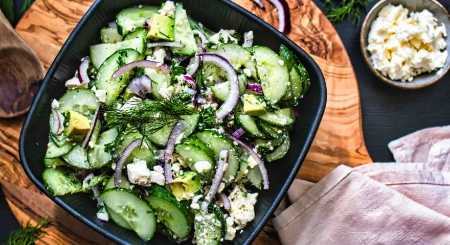 Erfrischender gurken feta salat mit Avocado, roten Zwiebeln und Dill, serviert in einer schwarzen Schüssel auf einem Holzbrett.
