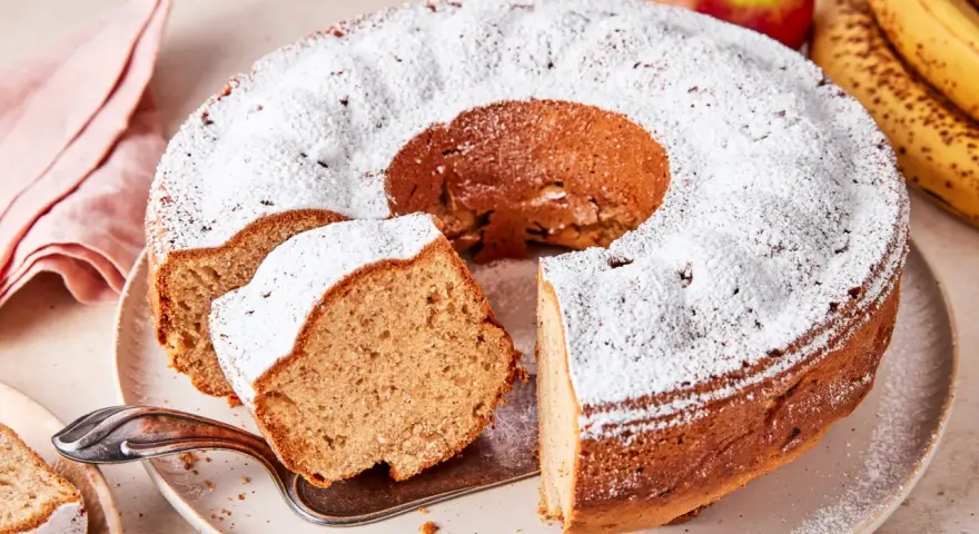 Saftiger Apfel-Bananen-Kuchen, bestäubt mit Puderzucker. Daneben liegen frische Äpfel und Bananen.