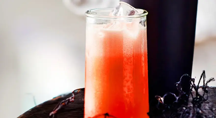 Erfrischender Aperol Sour mit Eiswürfeln in einem Glas, daneben ein dunkler Becher und Spinnweben-Deko.