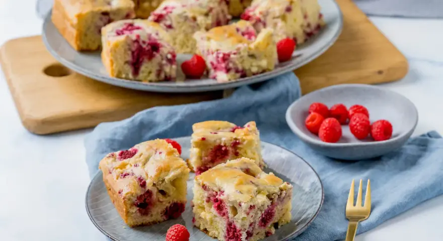 Saftiger Kuchen mit Himbeeren, auf Tellern und einer Platte angerichtet, daneben eine Schale mit frischen Himbeeren.
