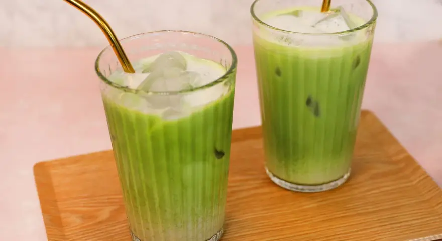 Zwei erfrischende Iced Matcha Latte mit goldenen Strohhalmen auf einem Holztablett. Perfekt für ein schnelles iced matcha latte rezept.
