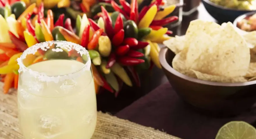 Ein erfrischendes Margarita-Rezept: Ein Glas mit Salzrand, Eiswürfeln und Limette, daneben bunte Chilis, Guacamole und Tortilla-Chips.