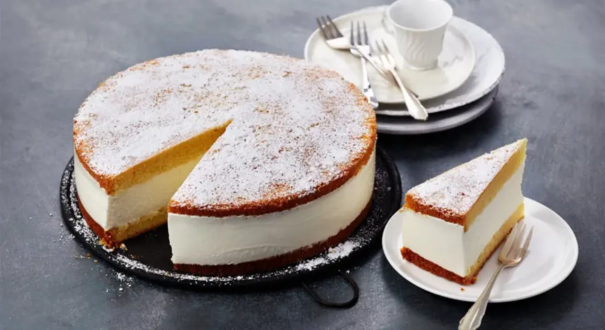 Eine köstliche Quark Sahne Torte, mit Puderzucker bestäubt, wartet auf ihre Genießer. Ein Stück ist bereits auf einem Teller angerichtet.