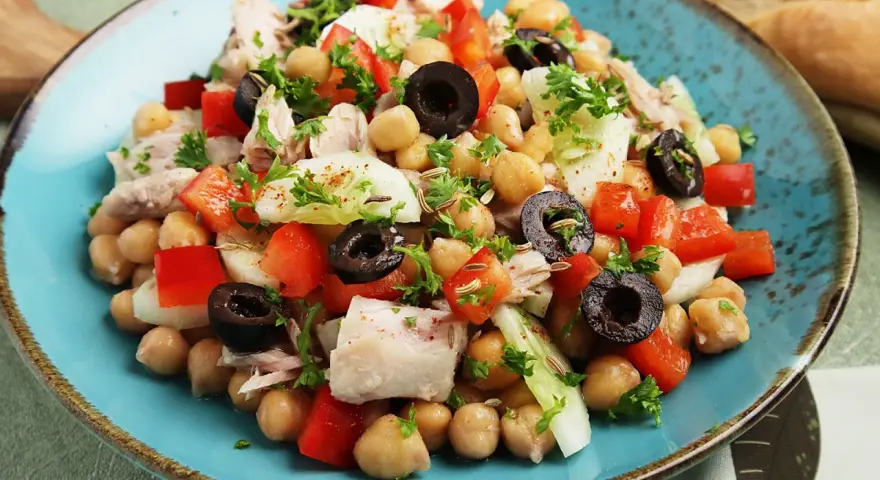 Ein bunter Kichererbsen Thunfisch Salat mit Paprika, Oliven und Petersilie, appetitlich auf einem blauen Teller angerichtet.