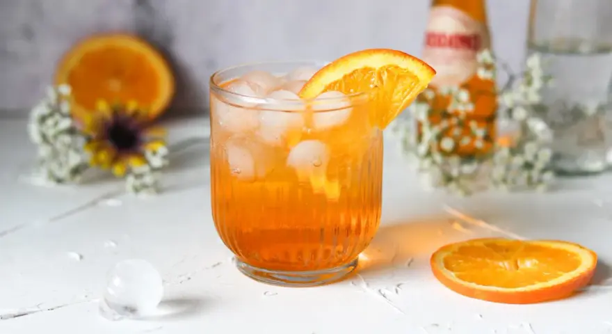 Erfrischender Crodino Spritz mit Eiswürfeln und Orangenscheibe, dekoriert mit kleinen weißen Blüten.