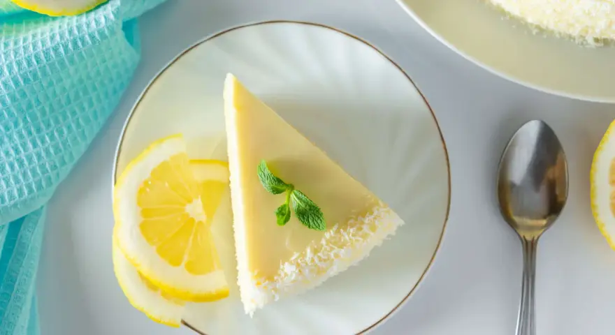 Ein Stück Philadelphia Torte Zitrone, garniert mit Minze und Kokosraspeln, neben Zitronenscheiben und einem Löffel.
