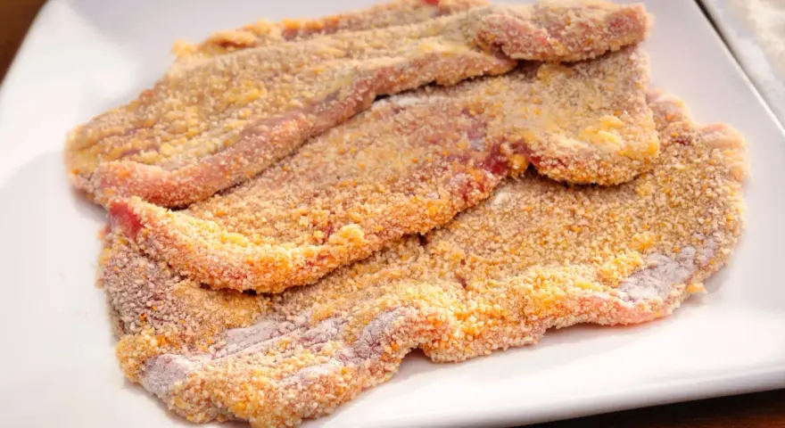Mehrere rohe Schnitzel liegen auf einem weißen Teller, bereit zum Schnitzel panieren. Die Panade ist goldbraun und knusprig.