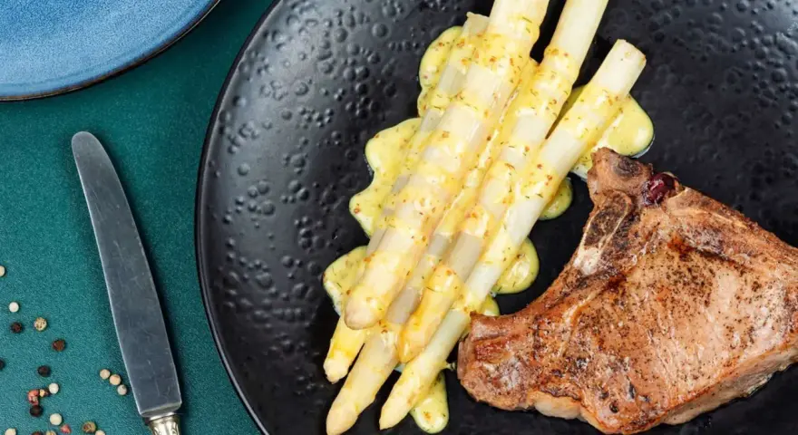Ein saftiges Schweinekotelett liegt neben einem Haufen cremigen Spargel in der Pfanne, beträufelt mit einer würzigen Sauce.