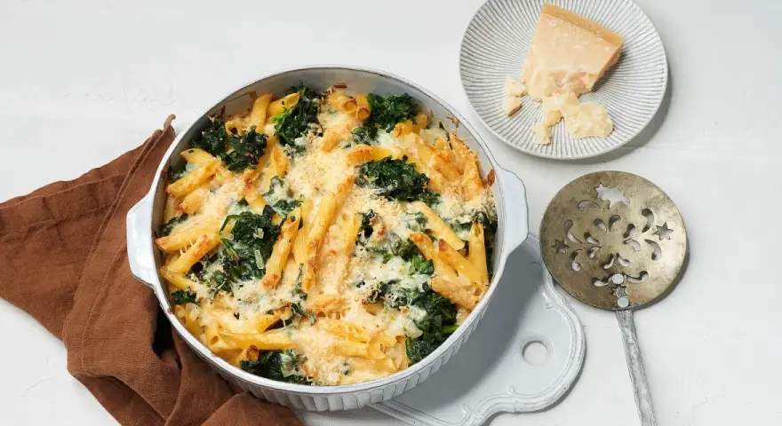 Herzhafter nudelauflauf mit spinat, überbacken mit Käse. Daneben ein Stück Parmesan und ein dekoratives Servierbesteck.