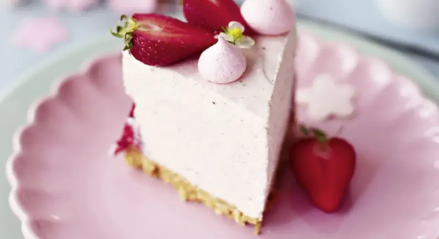 Ein Stück Erdbeer Cheesecake ohne Backen, dekoriert mit frischen Erdbeeren und rosa Baiser-Tupfern auf einem rosa Teller.