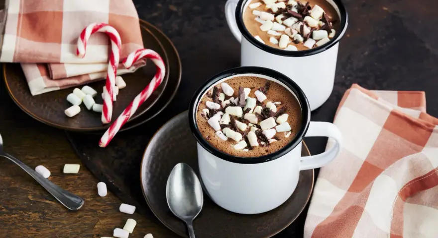 Zwei Tassen heißer Kakao mit Marshmallows und Schokostückchen, daneben Zuckerstangen und eine karierte Serviette.