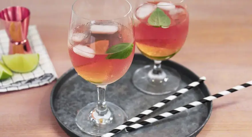 Zwei Gläser Rosato Spritz mit Eis und Minze, dazu Limettenspalten und ein Messbecher. Perfekt für einen sommerlichen Genuss.
