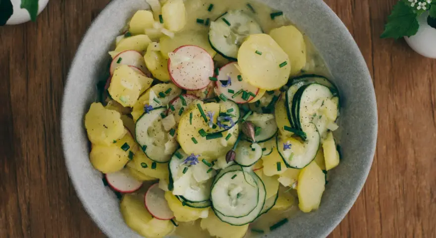 Ein Teller mit lauwarmer bayrischer Kartoffelsalat, garniert mit Radieschen, Gurken und Schnittlauch.