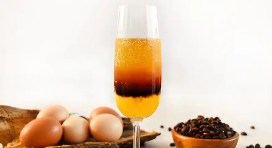 Schicht-Cocktail mit Ei, Kaffee und Orangensaft. Daneben liegen Eier und Kaffeebohnen.
