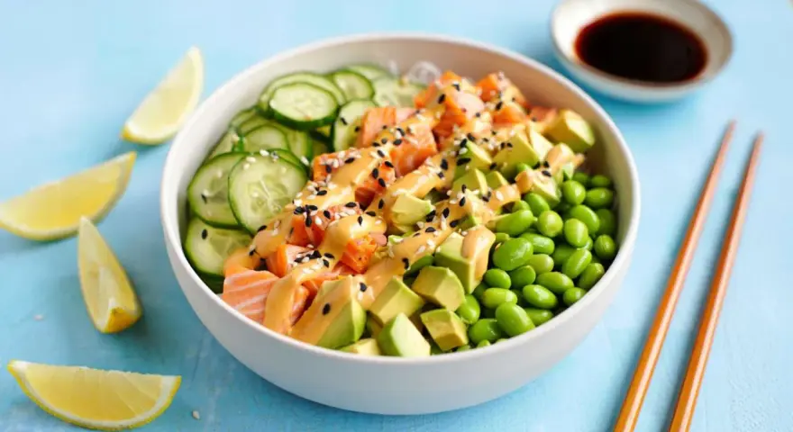 Schüssel mit einem frischen Salat aus Lachs, Avocado, Edamame und Gurkenscheiben, beträufelt mit cremiger Sauce und Sesam. Zitronenspalten und Sojasauce daneben.