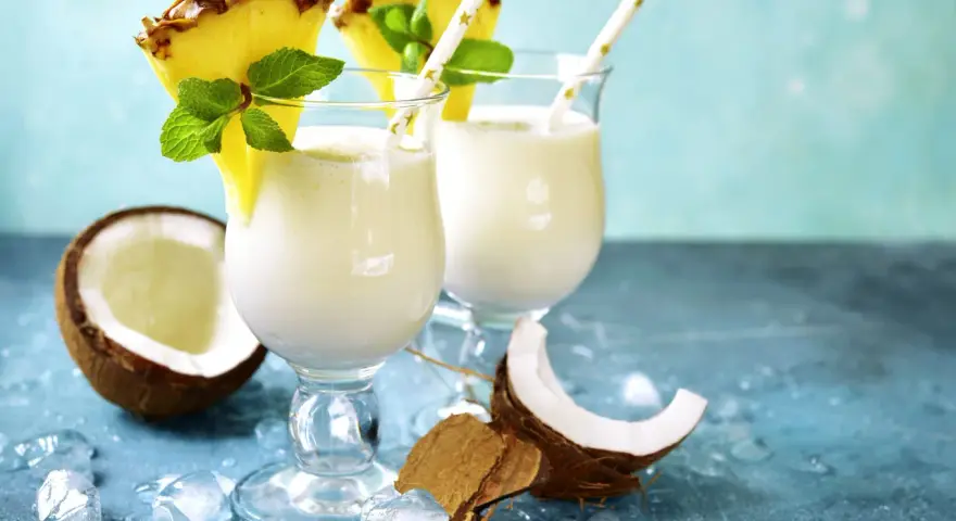 Zwei cremige Malibu Cocktails mit Ananas und Minze, umgeben von Kokosnuss und Eis.