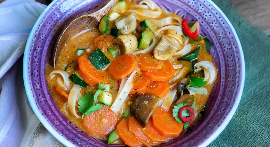 Eine dampfende, cremige Thai Curry Suppe mit Karotten, Zucchini, Pilzen und Reisnudeln in einer lila Schüssel.