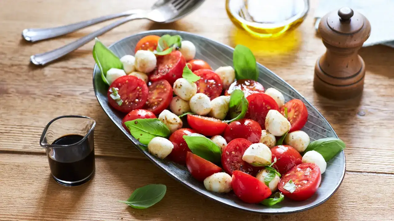 Ein frisches Rezept: Tomate Mozzarella Salat mit Basilikum, serviert auf einem ovalen Teller. Daneben ein kleines Glas Balsamico und ein Pfefferm&uuml;hlen.