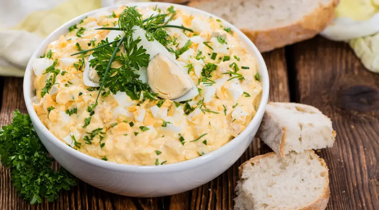 Cremiger Eiersalat mit Joghurt, garniert mit frischer Petersilie und Dill, serviert auf rustikalem Holz mit Brot.