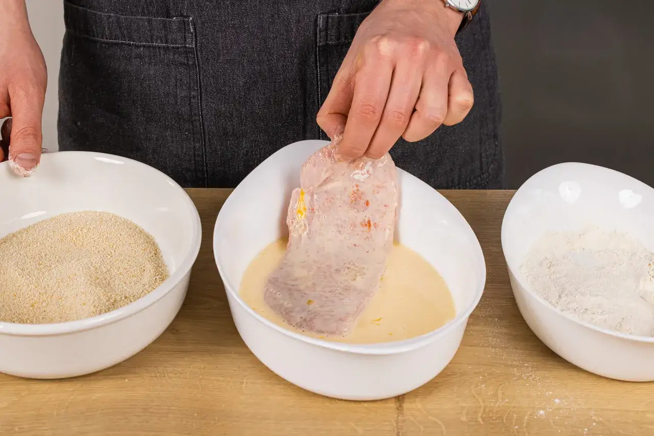 Schneiden wird in Ei getaucht, um es f&uuml;r das Schnitzel panieren vorzubereiten. Daneben stehen Sch&uuml;sseln mit Mehl und Paniermehl.