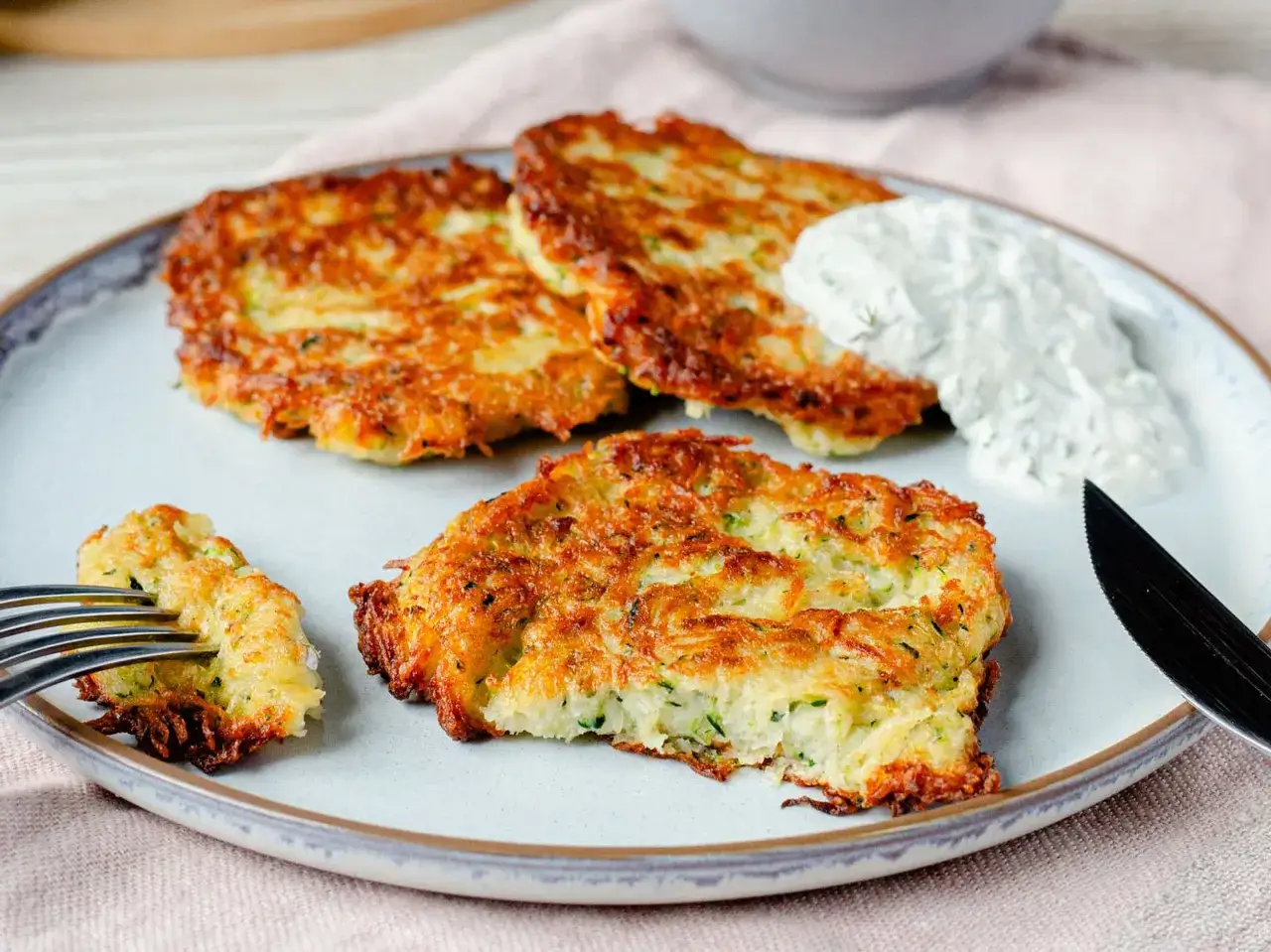 Goldbraune zucchini kartoffel puffer auf einem Teller, mit einem Klecks Dip und einer Gabel, die ein St&uuml;ck aufnimmt.