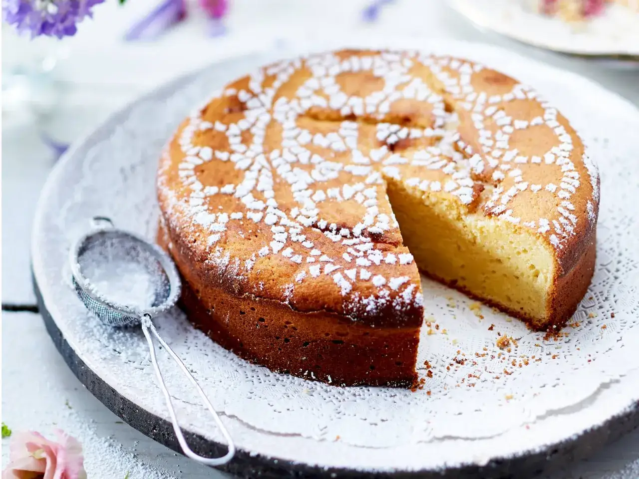 Ein saftiger Kuchen mit Joghurt, best&auml;ubt mit Puderzucker. Daneben ein Sieb, bereit f&uuml;r mehr S&uuml;&szlig;e.