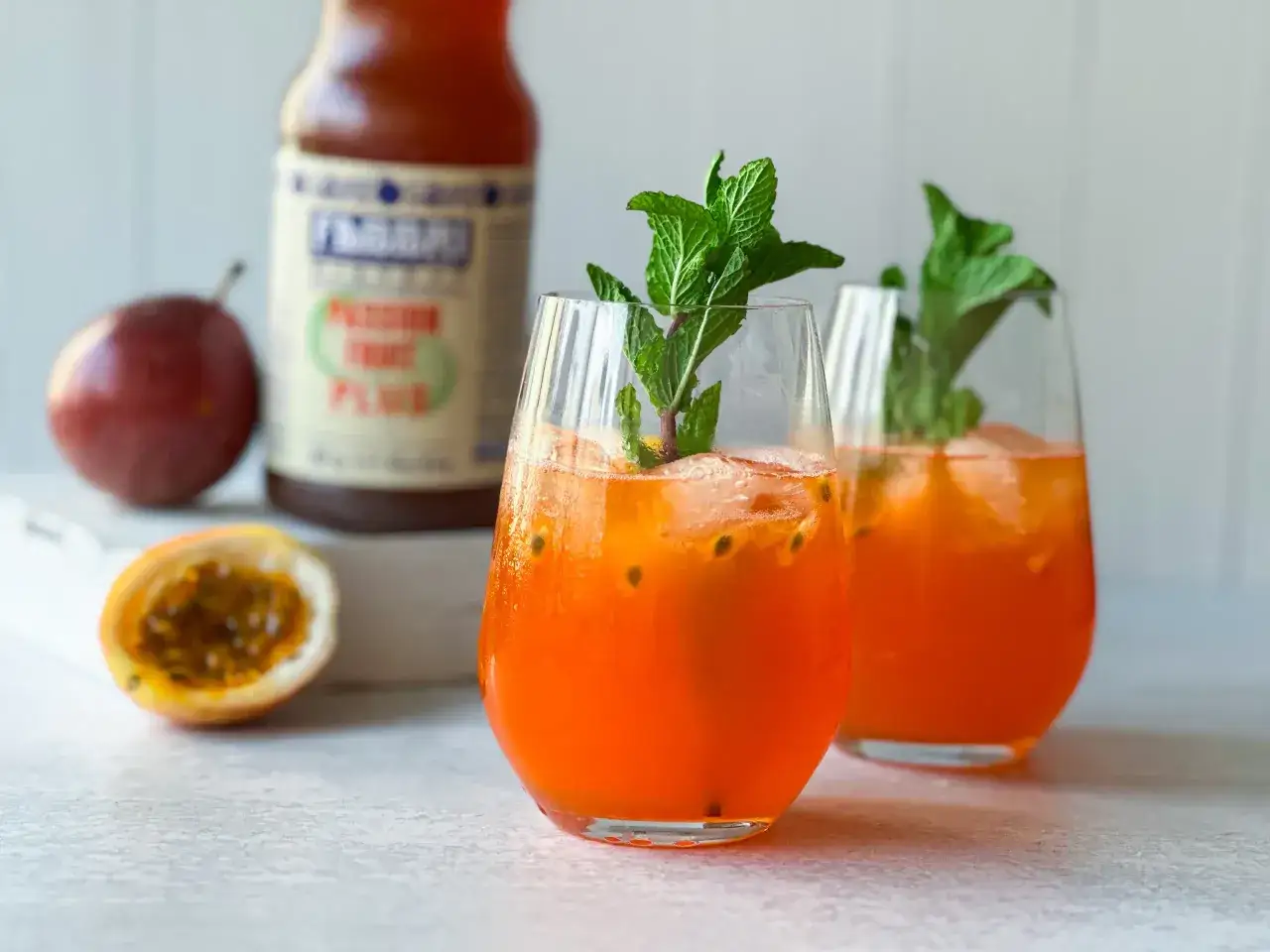 Zwei erfrischende Gl&auml;ser Maracuja Spritz mit Minze, Eis und Passionsfruchtst&uuml;cken. Eine Flasche im Hintergrund.