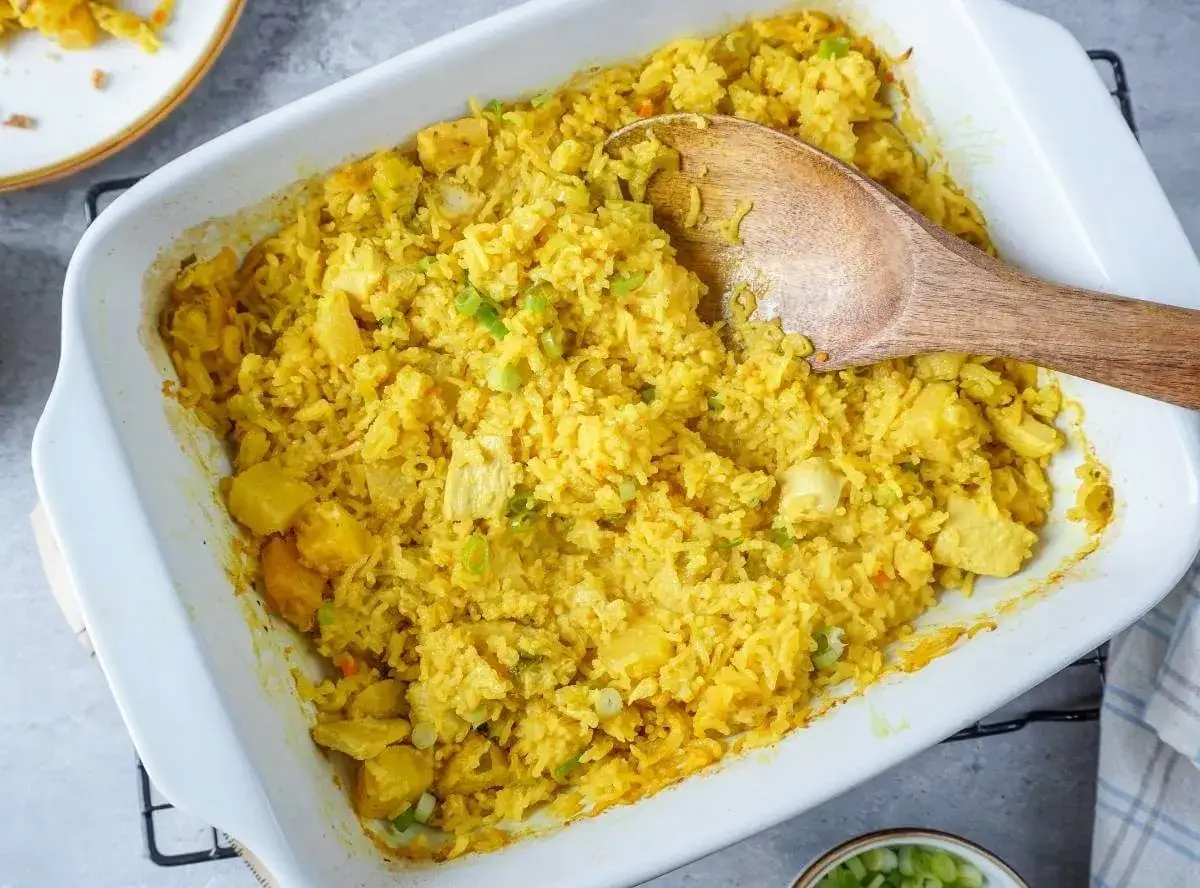 Ein gelber Reisauflauf mit Ananasst&uuml;cken und H&uuml;hnchen wird in einer wei&szlig;en Auflaufform serviert. Ein Holzl&ouml;ffel liegt darin. Perfekt, um Reis im Ofen zu kochen.