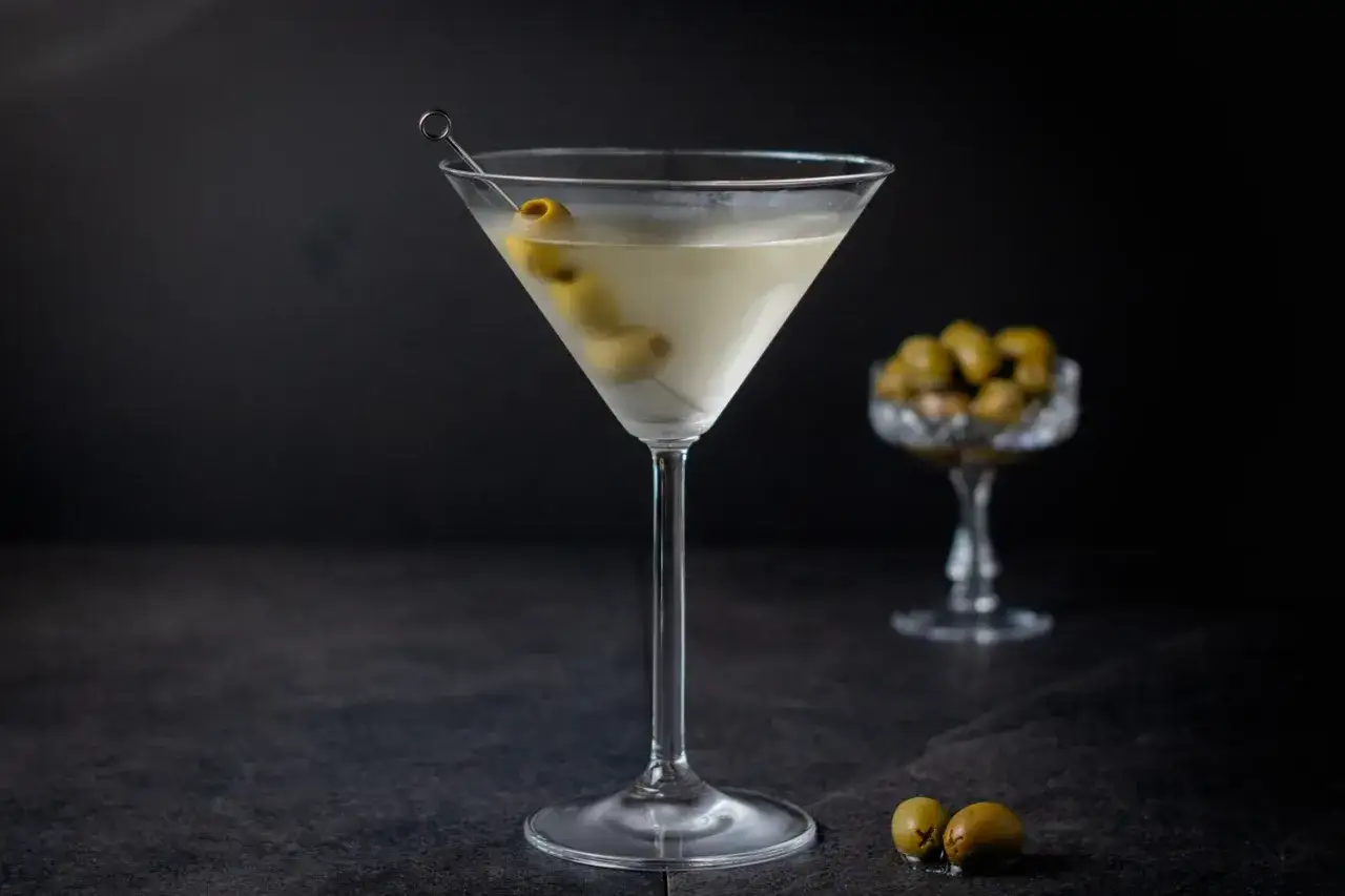Ein klassisches Martini-Rezept: Ein Martini-Glas mit einem Drink und Oliven, daneben eine Schale mit Oliven.