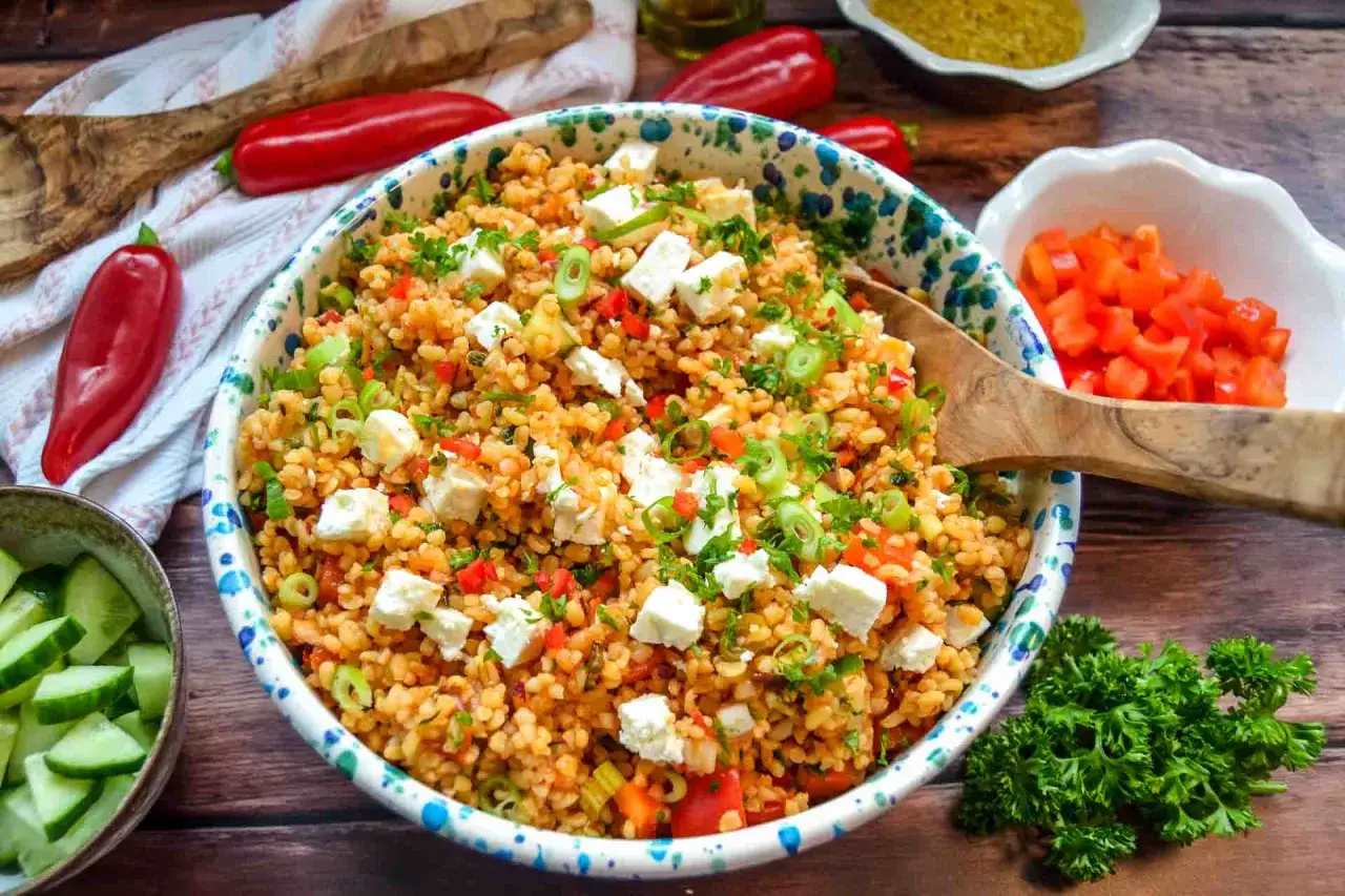 Frischer bulgursalat mit feta, Paprika und Kr&auml;utern, garniert mit Fr&uuml;hlingszwiebeln. Daneben liegen rote Peperoni und Gurkenscheiben.