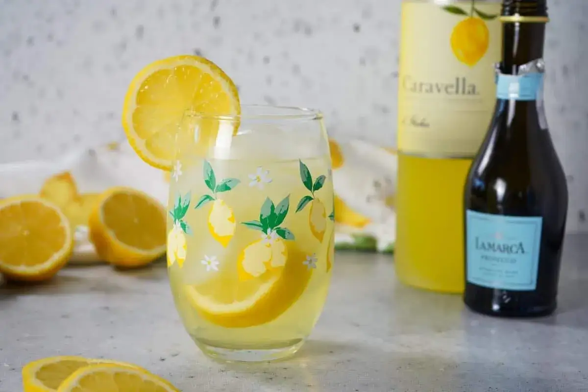 Erfrischender Limoncello Spritz im Glas mit Zitronenscheibe. Daneben Limoncello-Flasche und Prosecco.