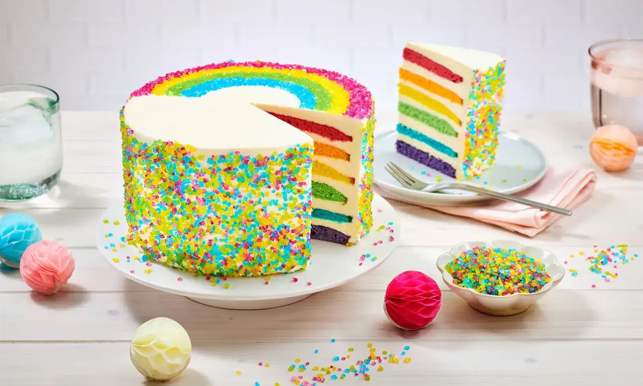 Bunter Geburtstagskuchen mit Regenbogenf&uuml;llung und Streuseln, perfekt f&uuml;r Kinder. Ein St&uuml;ck ist angeschnitten und zeigt die bunten Schichten.