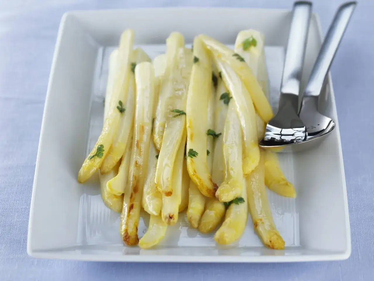 Zarter Spargel in der Pfanne, goldbraun gebraten und mit Petersilie garniert, liegt auf einem weißen Teller. Daneben liegen Besteckteile.