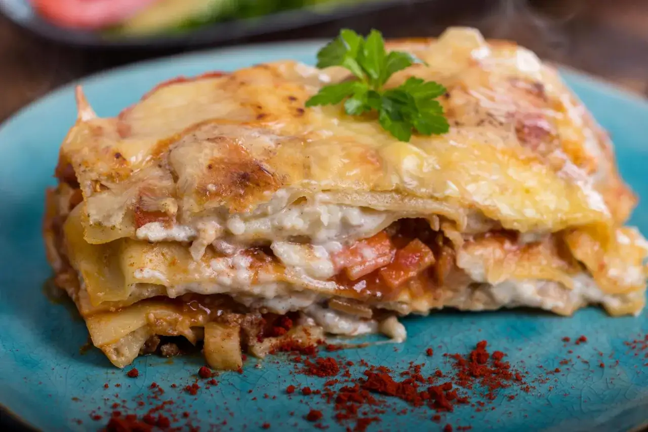 Ein Stück Lasagne mit Schmand, überbacken mit Käse und garniert mit Petersilie, auf einem blauen Teller.