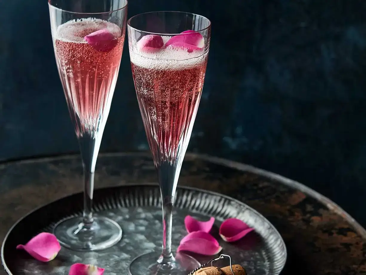 Zwei Gl&auml;ser mit einem rosafarbenen French 75, garniert mit Rosenbl&auml;ttern, auf einem Tablett.