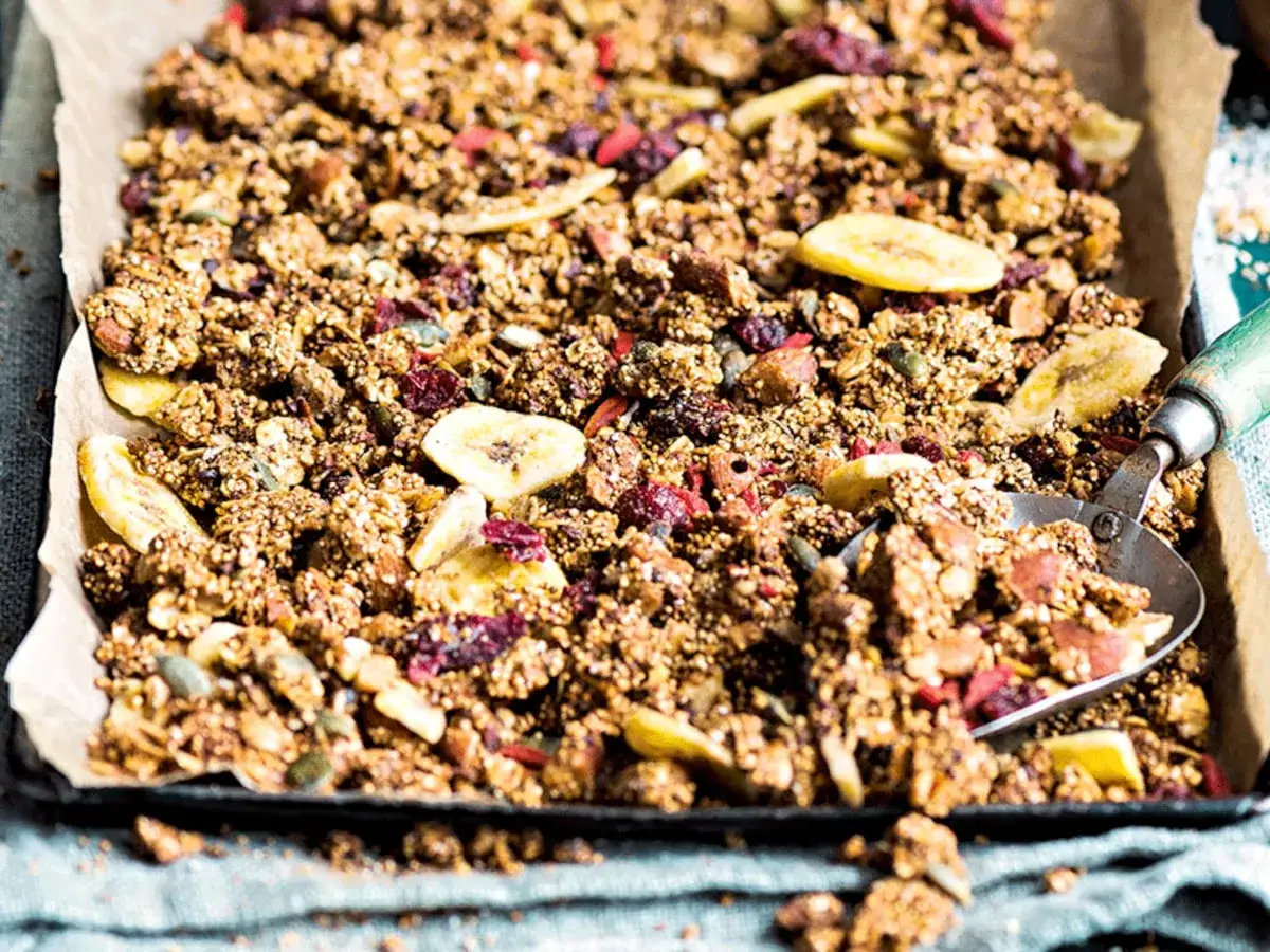 Knuspriges Granola mit Amaranth, Bananenchips und Cranberries auf Backpapier. Ein L&ouml;ffel nimmt eine Portion auf.