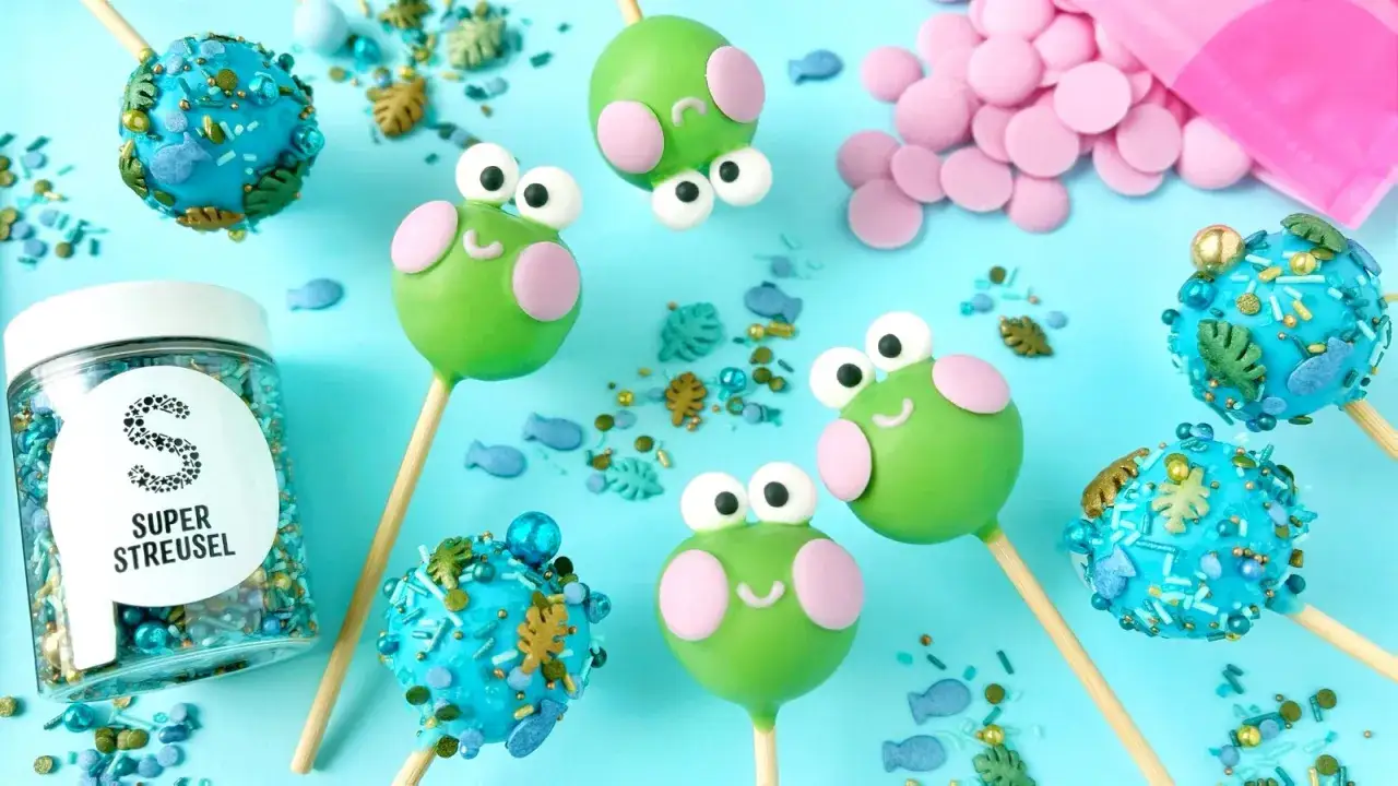 S&uuml;&szlig;e Frosch-Cake-Pops und blaue Ozean-Cake-Pops, inspiriert von einem Cake Pops Rezept. Streusel und rosa Schokolade liegen bereit.