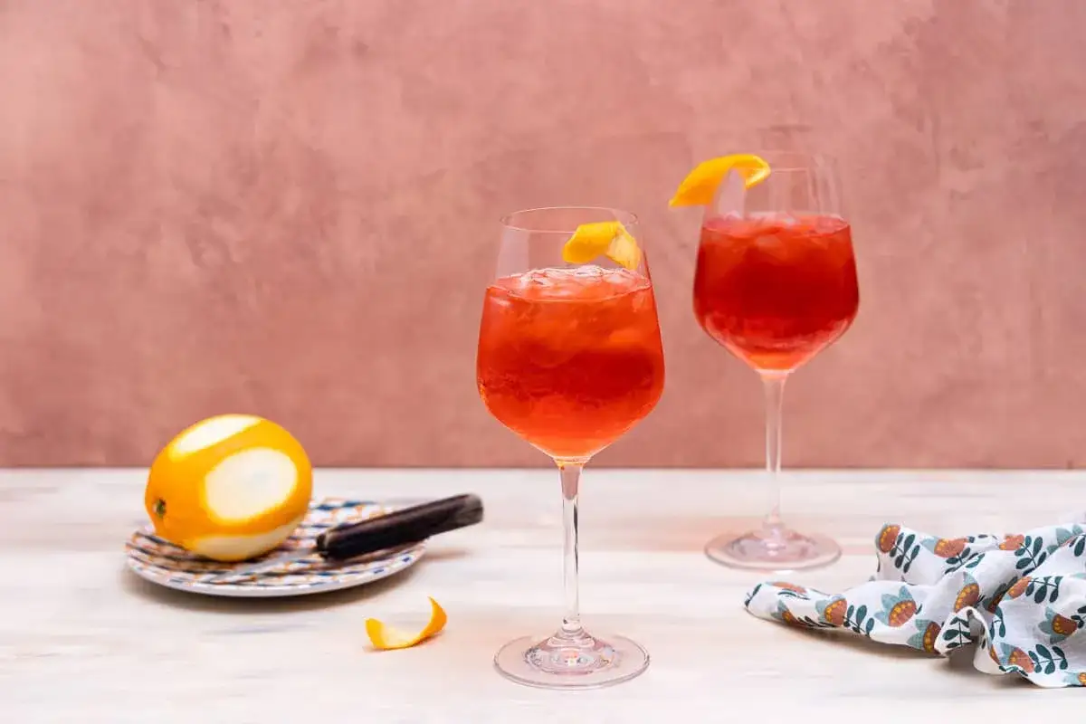Zwei erfrischende Campari Spritz mit Eis und Orangenzeste, daneben eine Orange und ein St&uuml;ck Zimtstange.