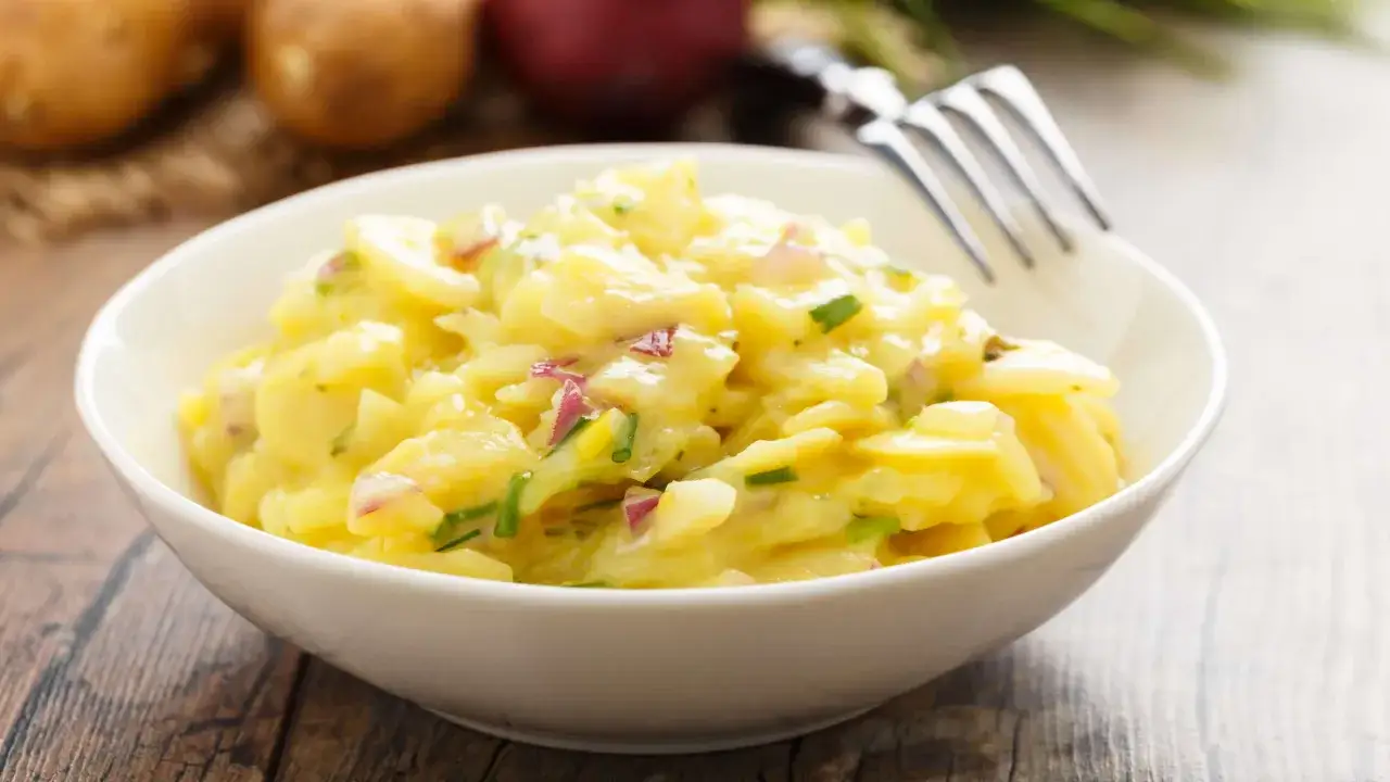 Ein cremiger Kartoffelsalat mit roten Zwiebeln und Schnittlauch, serviert in einer wei&szlig;en Sch&uuml;ssel. Perfekt f&uuml;r ein Picknick oder als Beilage.