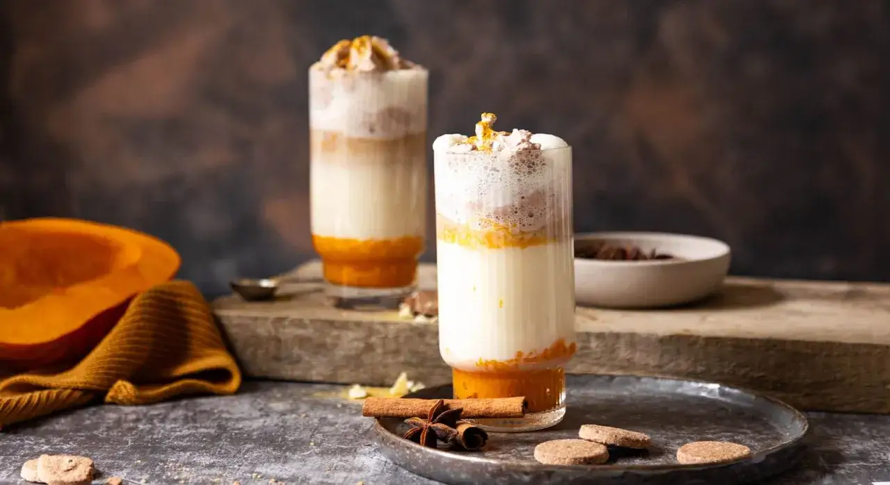 Zwei Gl&auml;ser mit geschichtetem Pumpkin Spice Latte ohne Kaffee, garniert mit Sahne und Zimt. Daneben eine halbe K&uuml;rbis.