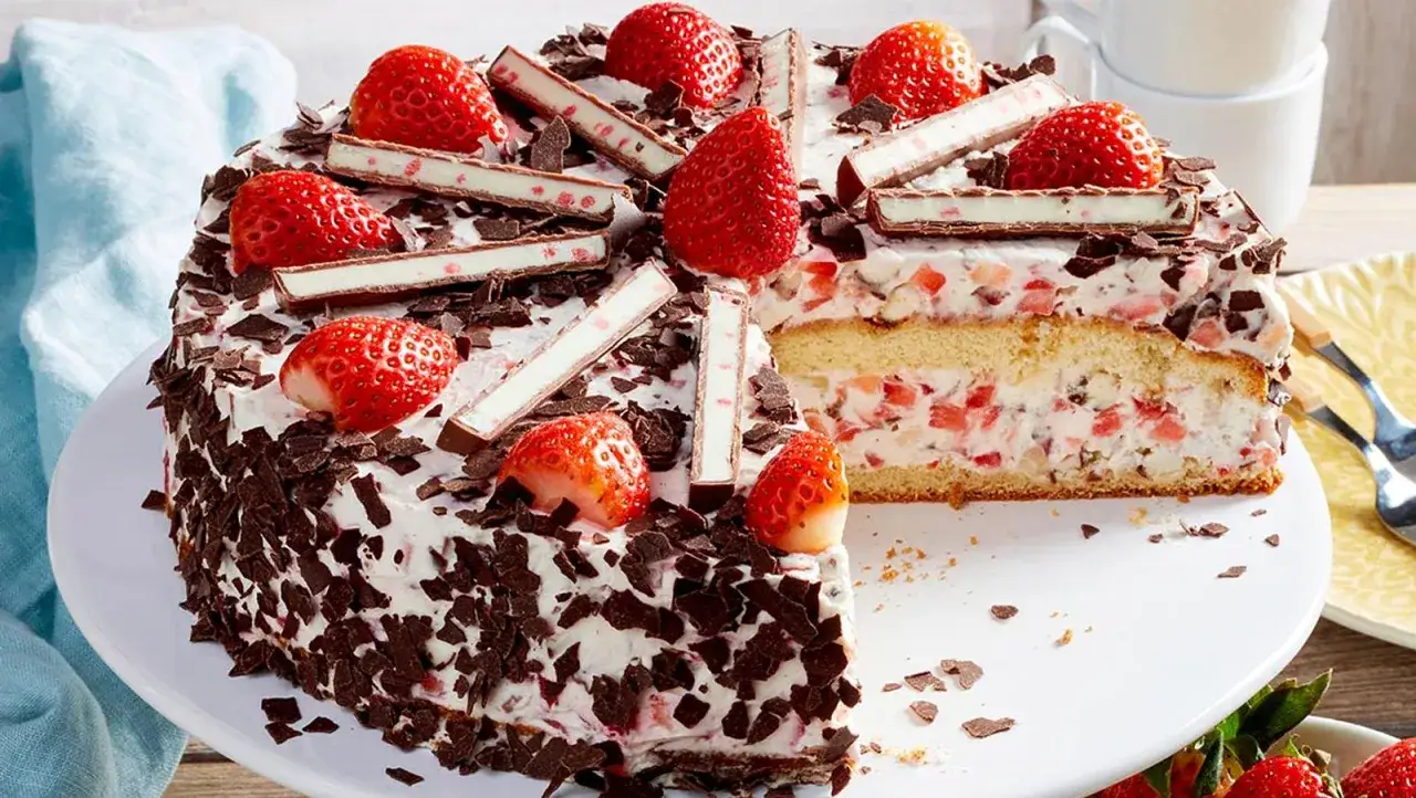 Erdbeer Yogurette Torte mit frischen Erdbeeren, Schokost&uuml;ckchen und wei&szlig;en Schokoriegeln dekoriert. Ein St&uuml;ck ist angeschnitten.