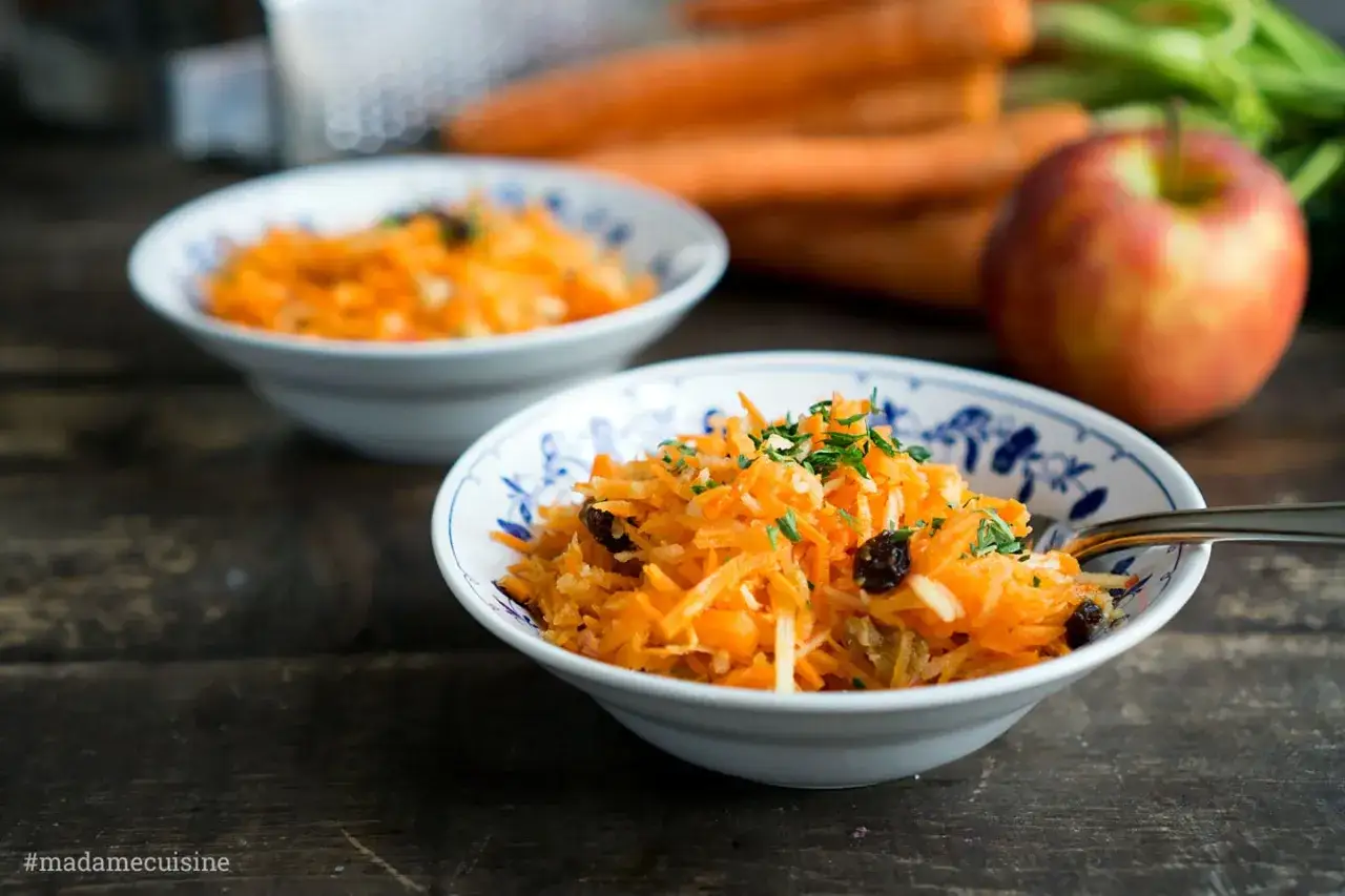 Zwei Sch&uuml;sseln mit Karotten-Apfel-Salat, garniert mit Petersilie und Rosinen. Im Hintergrund liegen frische Karotten und ein roter Apfel.