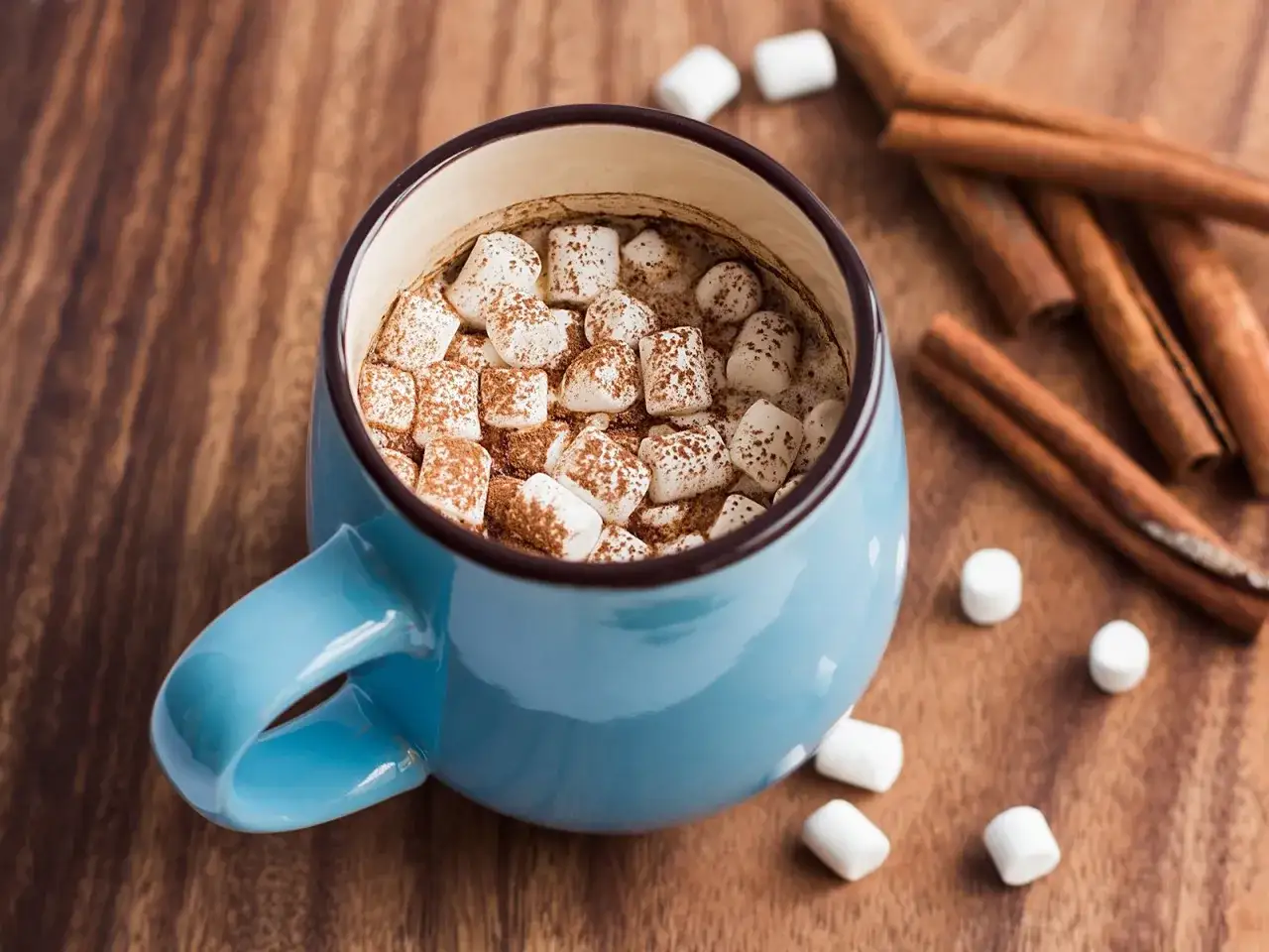Ein blauer Becher mit hei&szlig;em Kakao mit Marshmallows und Zimtstangen auf einem Holztisch.
