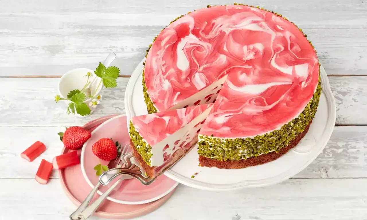 Eine wunderschöne Erdbeer Rhabarber Torte mit Marmor-Muster und Pistazienrand. Ein Stück wurde bereits angeschnitten.