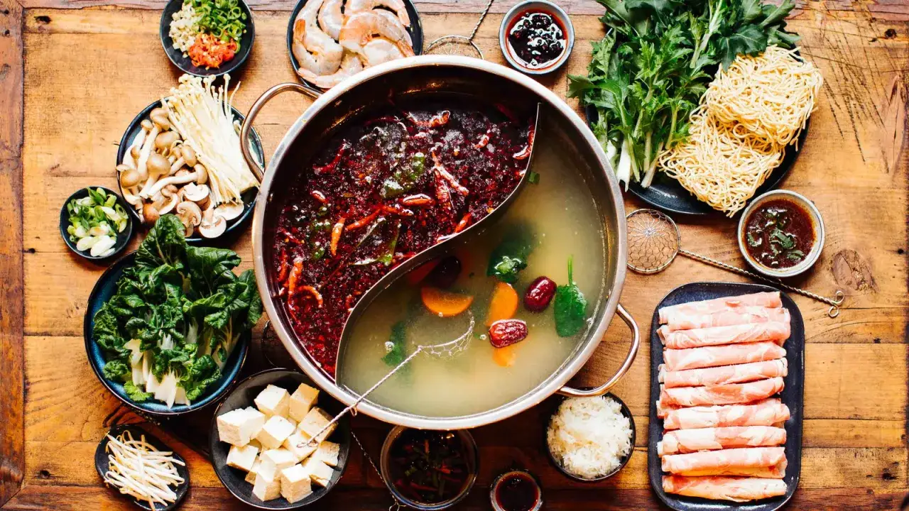 Alles für ein köstliches Hot Pot selber machen: Brühe, Fleisch, Gemüse, Tofu, Pilze, Garnelen, Nudeln und Dips auf Holztisch.